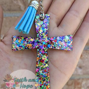 May include: A colorful glitter cross keychain with a light blue tassel. The cross is made of resin and has a variety of colors, including red, green, blue, and purple. The tassel is attached to a silver key ring.