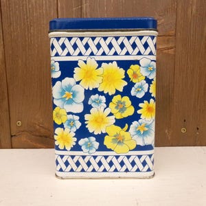 May include: A rectangular tin with a blue lid and a floral design. The tin features a pattern of yellow and blue flowers on a dark blue background. White geometric patterns border the top and bottom.