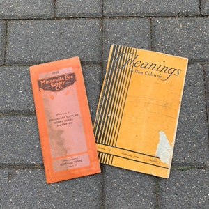 May include: Two vintage orange and yellow paperbacks on a gray concrete surface. The book on the left is titled "Minnesota Bee Supply Co." and the book on the right is titled "Meanings Bee Culture".