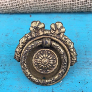 May include: A gold-toned metal drawer pull with a ring handle and ornate floral details. The pull has a circular design with a central flower motif.