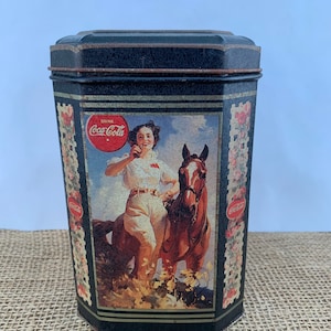 May include: A vintage black metal Coca-Cola tin with a colorful image of a woman riding a horse. The tin has a gold trim and the words "Drink Coca-Cola" are printed on the front.
