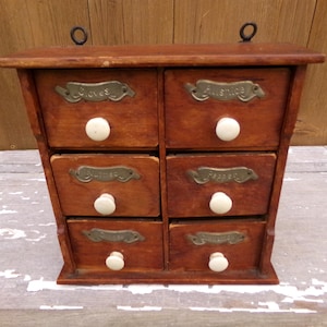 May include: A vintage wooden spice rack with six drawers. Each drawer has a white knob and a metal label. The labels read "Cloves", "Allspice", "Nutmeg", "Pepper", "Cinnamon", and "Ginger". The rack has two metal loops for hanging.