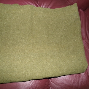 May include: An olive green blanket, folded and resting on a burgundy leather surface. The blanket has a textured appearance, suggesting a wool or similar material. The color is a muted, earthy tone.