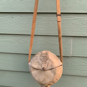 May include: A tan canvas bag with a brown strap. The bag has a flap with a button closure and a circular patch with the text "Boy Scouts of America National Council 100% Cotton".