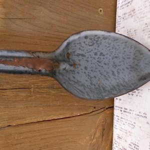 May include: Close-up of a vintage, gray enamelware spoon with a long handle. The spoon has a speckled surface and shows signs of rust. The spoon is resting on a wooden surface.