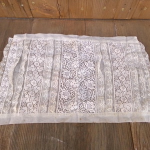 May include: A rectangular, off-white lace curtain or valance. The lace features floral and geometric patterns, with vertical bands of floral designs and a central panel of intricate lace. The edges are scalloped, and the fabric appears delicate.