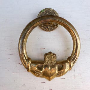 May include: A circular brass door knocker featuring a Claddagh design with two hands holding a heart topped by a crown. The metal has a weathered, golden patina and is attached to a small, round mounting plate with an intricate Celtic knot pattern.