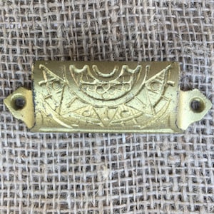 May include: Gold-toned metal drawer pull with an ornate design. The pull has two holes for mounting.