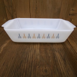 May include: A rectangular white Pyrex baking dish with handles. The dish is decorated with a repeating pattern of blue and orange starburst designs along the side. The dish is sitting on a wooden surface.