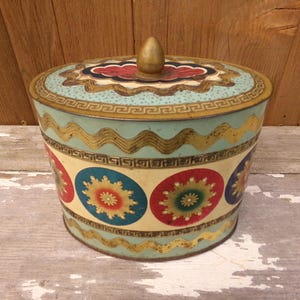 May include: Vintage round tin with a lid, featuring a light blue background with gold and cream-colored accents. The tin has a gold finial and is decorated with colorful floral designs. The tin is likely used for storage.