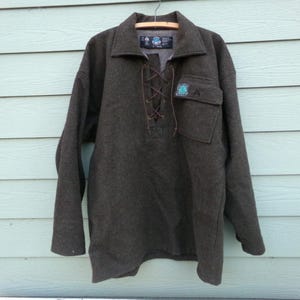 May include: A dark gray wool pullover shirt with a collared neckline and a lace-up closure. It features a pocket on the chest and a small patch with a logo. The shirt is hanging on a wooden hanger against a light blue wall.