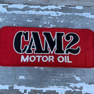 May include: Red rectangular patch with white stitching around the edges. The patch has white text that reads "CAM2 MOTOR OIL" with black lettering in a stylized font.