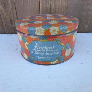 May include: Vintage round metal container with a floral design in orange, blue, and white. The container has a lid and features text that reads "Florient Flowers of the Orient Dusting Powder" and "Colgate & Co. New York U.S.A."