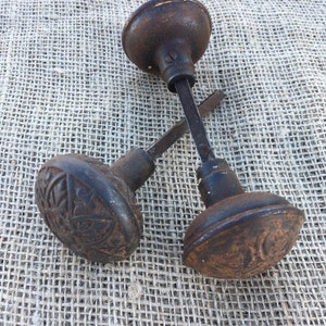 May include: Two antique cast iron door knobs with intricate designs. The knobs are rusty and have a dark brown patina. They are on a burlap background.