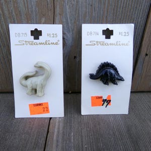 May include: Two dinosaur-shaped pins on white cards. One is a light beige Brontosaurus, the other is a dark blue Stegosaurus. Each card has the text "Streamline" and a clearance sticker.