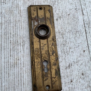 May include: A vintage brass door plate with a keyhole and a round knob. The plate has a worn, aged look with a patina finish.