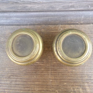May include: Two antique brass door knobs with a circular design and a raised rim. The knobs are on a wooden surface.