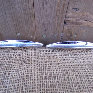 May include: Two polished, silver-colored drawer pulls with a streamlined, oval design. The pulls have a reflective surface and a dark, recessed center. They are displayed against a rustic wooden background and a burlap-like fabric.