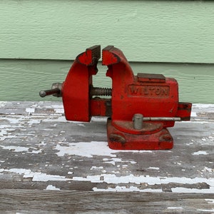 May include: A red Wilton brand metal vise with a silver handle. The vise is sitting on a weathered wooden surface.