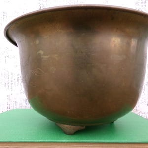 May include: A large, round, brass planter with a wide, flared rim and three short legs. The planter has a smooth, polished surface and appears to be vintage.