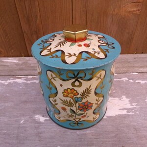 May include: A vintage, round, blue metal container with a lid. The container features a floral design with gold ribbon accents. The lid has a gold knob. The container is likely used for storage.