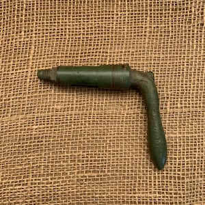 May include: An antique, dark green metal tool with a handle. The tool has a threaded end and a right-angle handle. The handle has a textured grip. The tool is set against a burlap background.