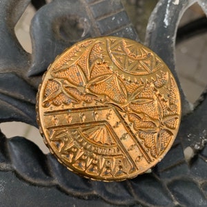May include: A round, gold-colored metal trinket box with an intricate, embossed floral design. The box has a raised, geometric pattern around the edge.