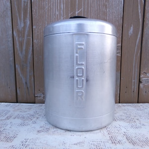 May include: A silver metal flour canister with a black lid and the word "FLOUR" embossed on the front.