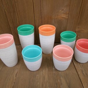 May include: A collection of seven cylindrical tumblers with a speckled white base and contrasting solid-colored rims in shades of coral, orange, and teal. The cups feature a subtle horizontal ribbed texture along the exterior of the white body.