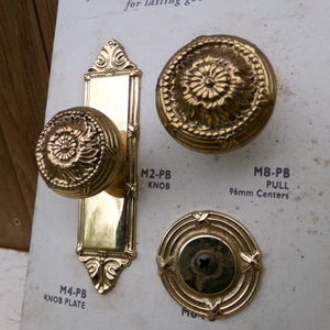 May include: Close-up of antique brass door hardware, including knobs and a pull. The knobs and pull have ornate floral designs. The text on the display reads "M4-PB KNOB PLATE", "M2-PB KNOB", and "M8-PB PULL 96mm Centers".