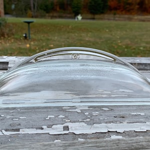 May include: A clear, curved glass lid with a handle and a small logo. The lid has a rectangular base and a rounded top. The background shows a blurred outdoor scene with trees and a bird bath.