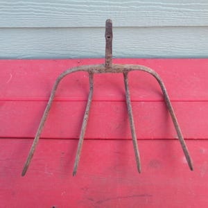 May include: An antique, rusty metal garden fork with four tines and a vertical handle. The tool is set against a red wooden surface with a light blue background, suggesting outdoor use and a vintage aesthetic.