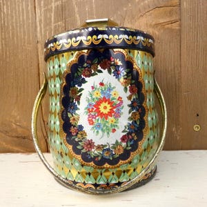 May include: Vintage metal container with a floral design. The container has a lid and a handle. The body of the container is decorated with a diamond pattern in green and gold. A central oval features a bouquet of colorful flowers.