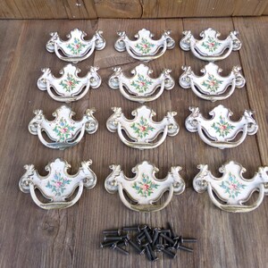 May include: Twelve white porcelain cabinet knobs with floral designs and gold accents. The knobs are arranged in three rows of four. Each knob has a ring pull handle. The knobs are accompanied by a pile of screws.