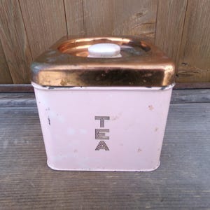 May include: A vintage pink metal tea canister with a copper lid and the word "TEA" printed on the front.