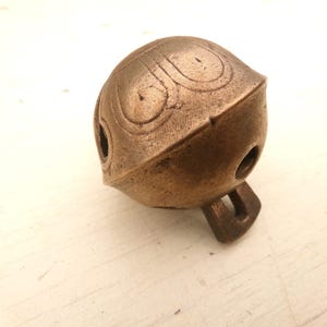 May include: A bronze-colored bell with a rounded top and a flat bottom. The bell has decorative swirl patterns on the top and a small loop for hanging. It has three holes and a rectangular loop at the bottom.