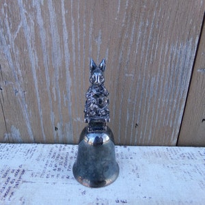 May include: A silver bell with a rabbit figurine on top. The rabbit is sitting on a base and holding a flower. The bell is on a white surface.