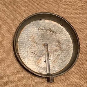 May include: A vintage Bake-King brand cake pan with a removable bottom. The pan is made of metal and has a silver finish. The pan is slightly rusted and has some scratches. The pan is sitting on a burlap surface.