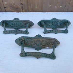 May include: Three antique bronze drawer pulls with a floral design. Each pull has a rectangular backplate with a row of flowers and a curved handle. The pulls are a weathered green color, suggesting age and use. They are placed on a white surface.