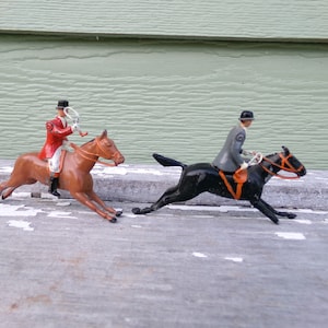 May include: Two toy horse riders, one in a red jacket and one in a gray jacket, are riding their horses. The horses are running, and the riders are holding their reins.