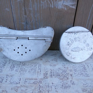 May include: Two vintage metal fishing tackle boxes. One is a round box with the words "Leader Box" printed on the lid. The other is a half-circle box with a hinged lid and a series of holes in the bottom.