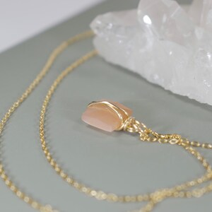 Pu&ograve; includere: Una collana con catena color oro con un ciondolo quadrato color pesca. Il ciondolo &egrave; avvolto in filo d'oro. La collana &egrave; esposta su una superficie grigia con una formazione di cristallo bianco sullo sfondo.