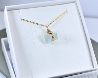 Può includere: Una delicata collana a catena dorata con un ciondolo con due pietre di acquamarina blu chiaro avvolte in filo d'oro. La collana è esposta in una scatola bianca.