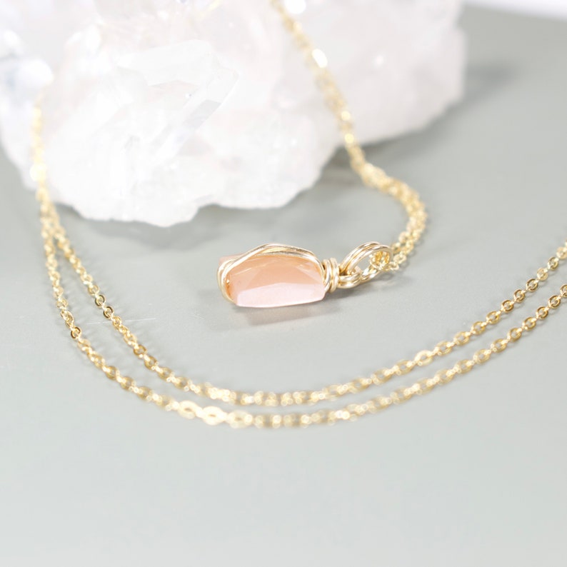 Pu&ograve; includere: Una delicata collana color oro con un pendente in pietra preziosa color pesca. Il pendente &egrave; avvolto in filo metallico e appeso a una sottile catena. La collana &egrave; esposta su una superficie grigio chiaro, mettendo in risalto il suo design elegante.