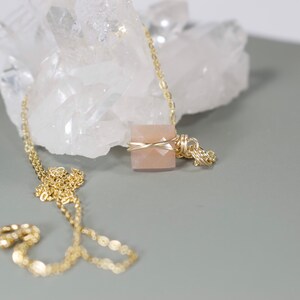 Pu&ograve; includere: Una collana dorata con un pendente quadrato in pietra preziosa color pesca. Il pendente &egrave; avvolto in filo d'oro e appeso a una delicata catena d'oro. Lo sfondo include un cristallo bianco e una superficie grigia.