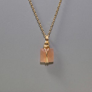 Pu&ograve; includere: Una collana a catena color oro con un ciondolo quadrato color pesca. Il ciondolo &egrave; avvolto in filo metallico color oro. La collana &egrave; esposta su uno sfondo grigio.