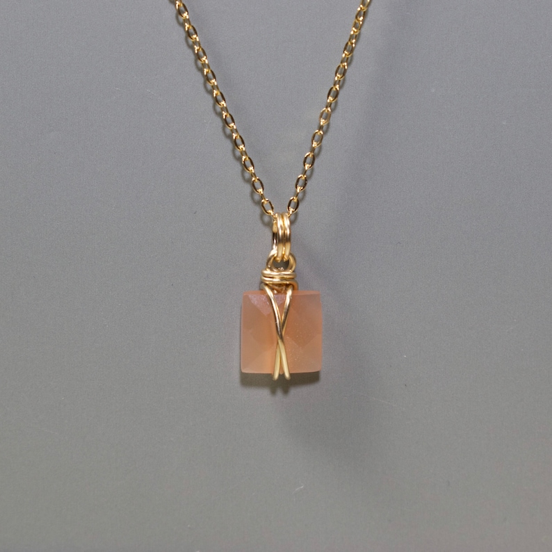 Pu&ograve; includere: Una collana a catena color oro con un ciondolo quadrato color pesca. Il ciondolo &egrave; avvolto in filo metallico color oro. La collana &egrave; esposta su uno sfondo grigio.