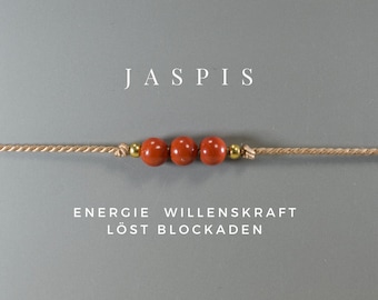 Può includere: Un braccialetto di perline di diaspro rosso con accenti dorati su uno stringa beige. Il testo "JASPIS" è sopra il braccialetto e il testo "ENERGIE WILLENKRAFT LÖST BLOCKADEN" è sotto il braccialetto.