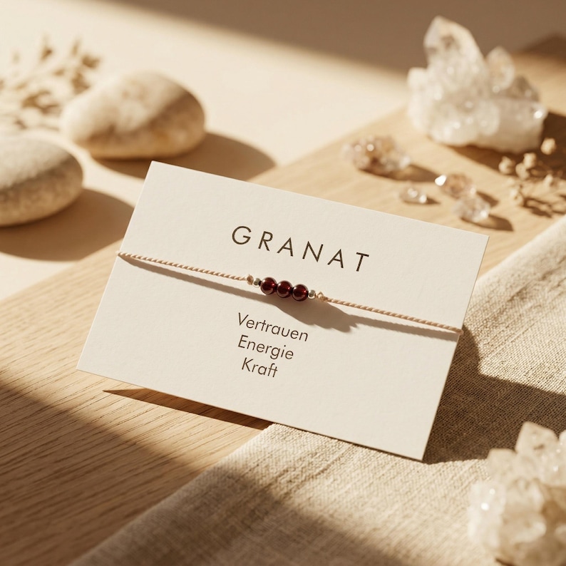 May include: A fine bracelet with three deep red garnet beads on a thin, beige cord. The bracelet is presented on a card with the word "GRANAT" and German words "Vertrauen, Energie, Kraft". The background includes crystals and smooth stones.