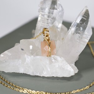 Pu&ograve; includere: Una collana color oro con un ciondolo a forma di pietra preziosa quadrata color pesca. La pietra preziosa &egrave; avvolta in filo d'oro e pende da una delicata catena d'oro. La collana &egrave; esposta su una formazione di cristallo bianco.